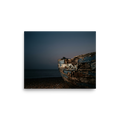 'The Faded Blue Boat' — a Art photography print. Close-up photograph of a weathered wooden boat with peeling blue paint resting on a rocky beach at dusk, suitable for a serene bathroom or living room.