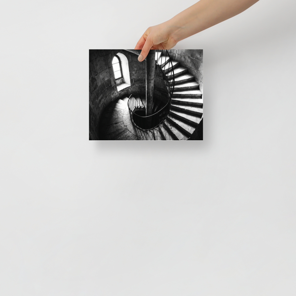 'Descent into Light' — a Art photography print. A high-contrast black and white photograph looking down a spiral stone staircase illuminated by a small window, adding depth and intrigue to an Office or Hallway.