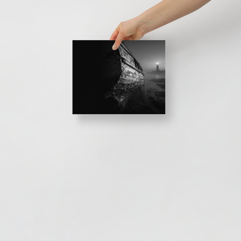 'Final Anchor' — a Art photography print. Black and white photograph of an old, peeling wooden boat resting on rocks with a glowing lighthouse in the foggy distance, perfect for a cozy living room.