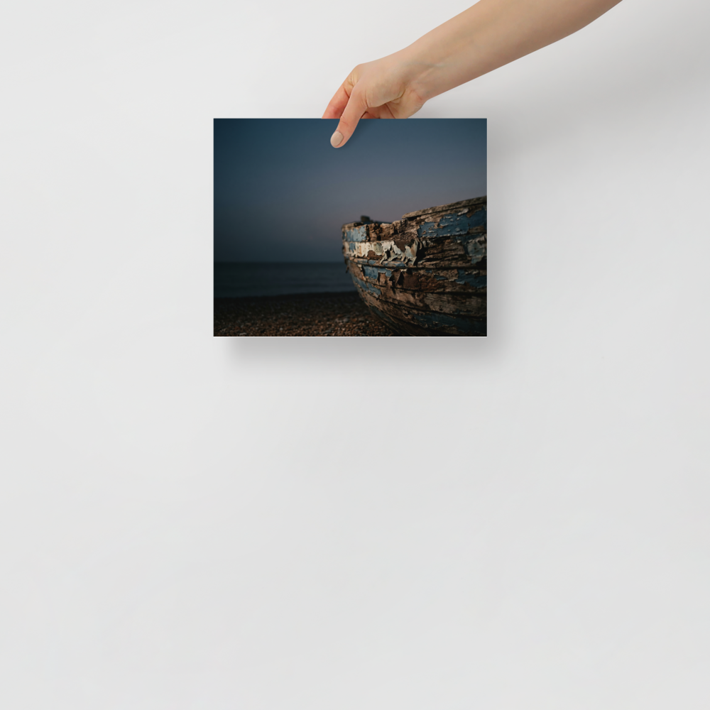 'The Faded Blue Boat' — a Art photography print. Close-up photograph of a weathered wooden boat with peeling blue paint resting on a rocky beach at dusk, suitable for a serene bathroom or living room.