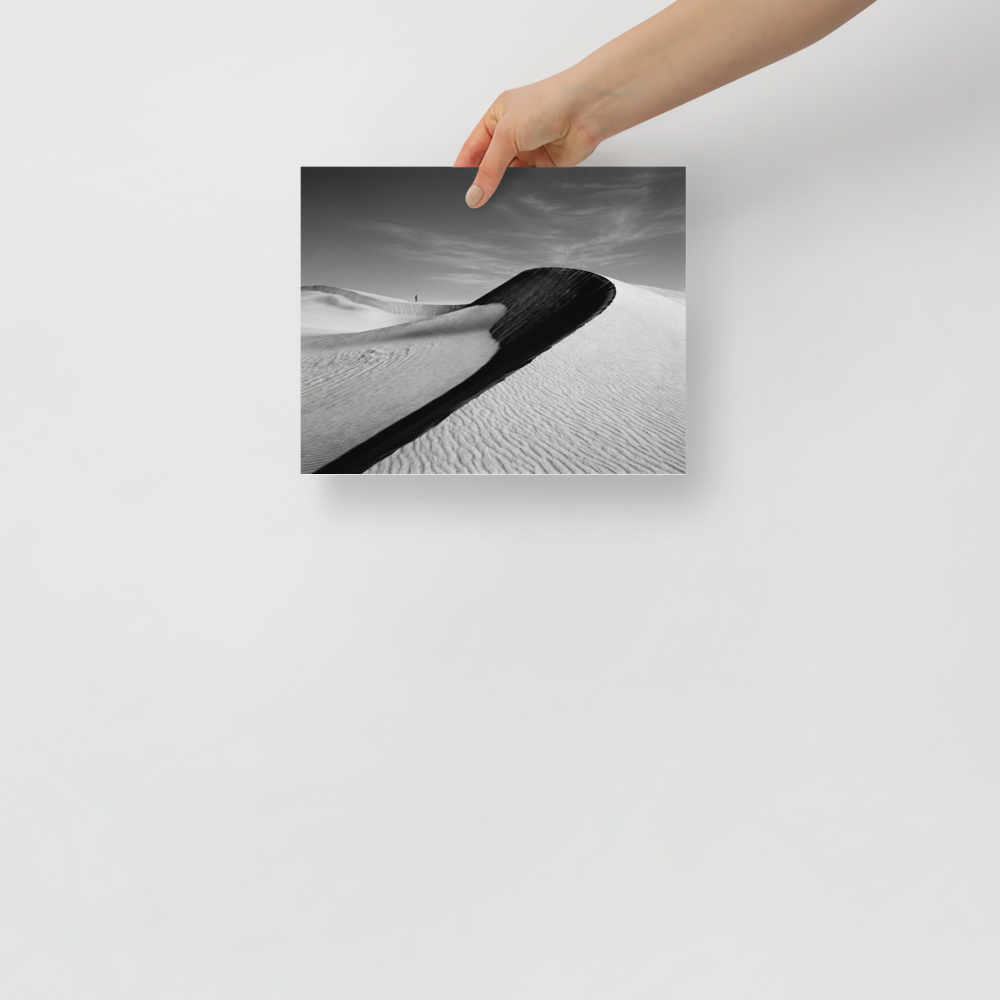 'Footsteps in the Void' — a Art photography print. A minimalist black and white photograph of sweeping sand dunes with a tiny figure walking along the ridge, perfect for adding a sense of scale to an Office.