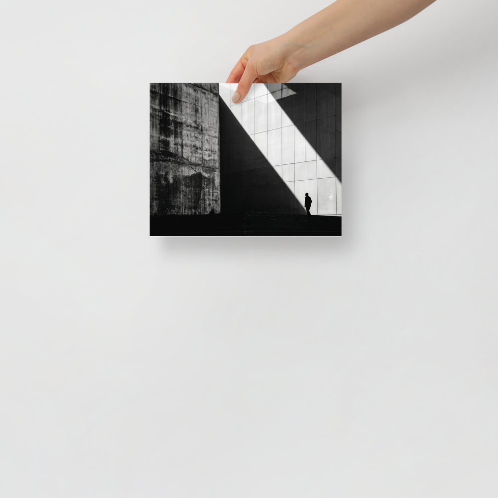 'Urban Geometry' — a Modernism print. A high-contrast black and white architectural photograph featuring a sharp diagonal shaft of light and a lone silhouette walking past textured concrete walls, perfect for an Office or Modern Living Room.