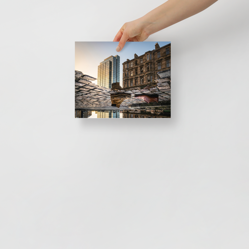 'Steps Through Time' — a Art photography print. Creative puddle reflection photograph showing a work boot and a dress shoe on cobblestones, with a modern glass skyscraper and an old stone building in the background, ideal for an office or hallway.
