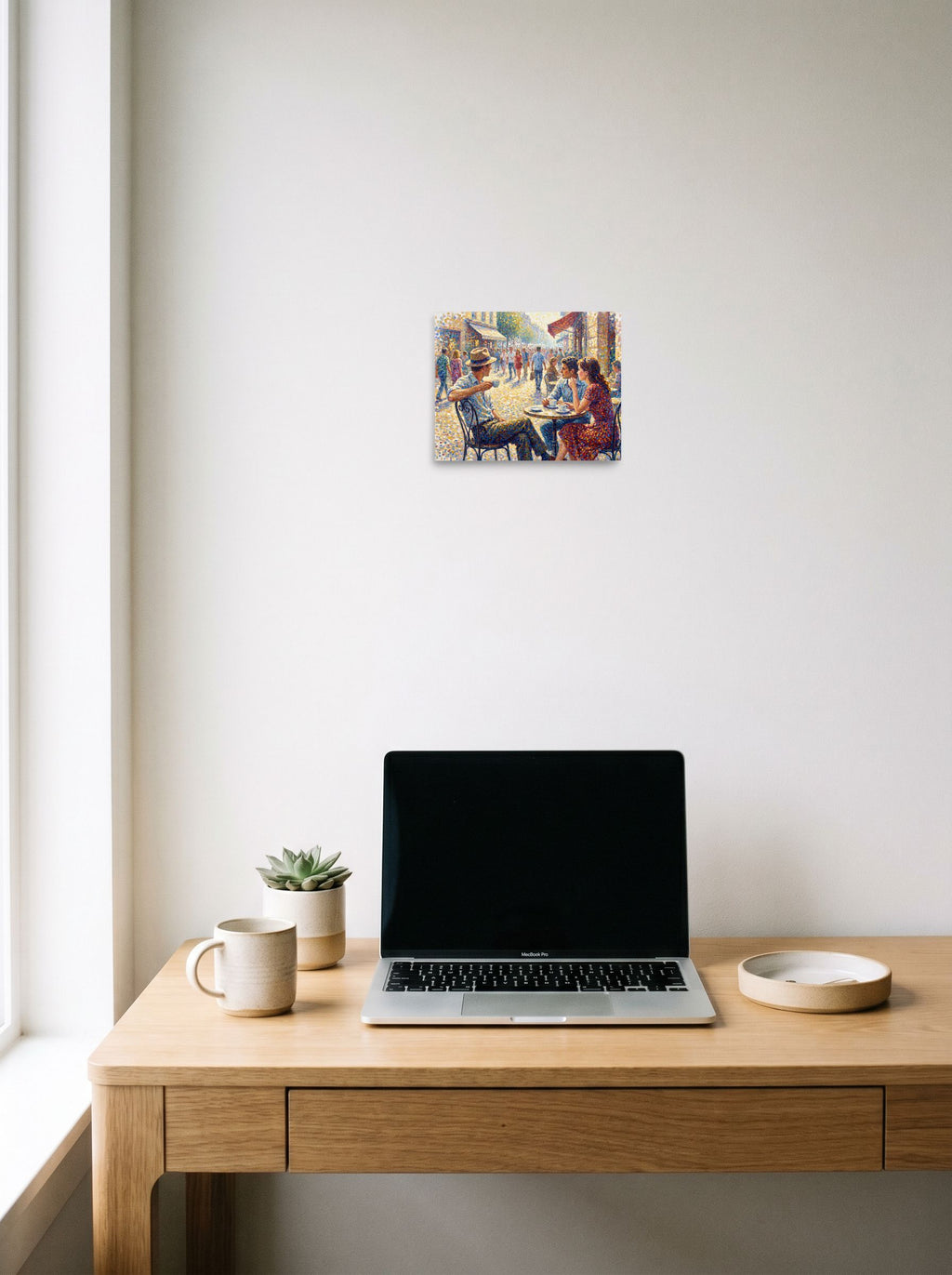 A unframed paper print of the Pointillism artwork 'Midday at the Bistro', hanging above a worktable with a laptop.