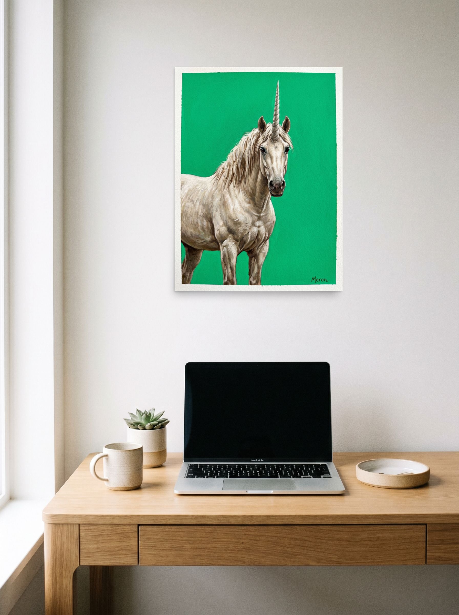 A 18x24 inch unframed paper Realism print of 'The Emerald Unicorn' hanging above a worktable with a laptop.