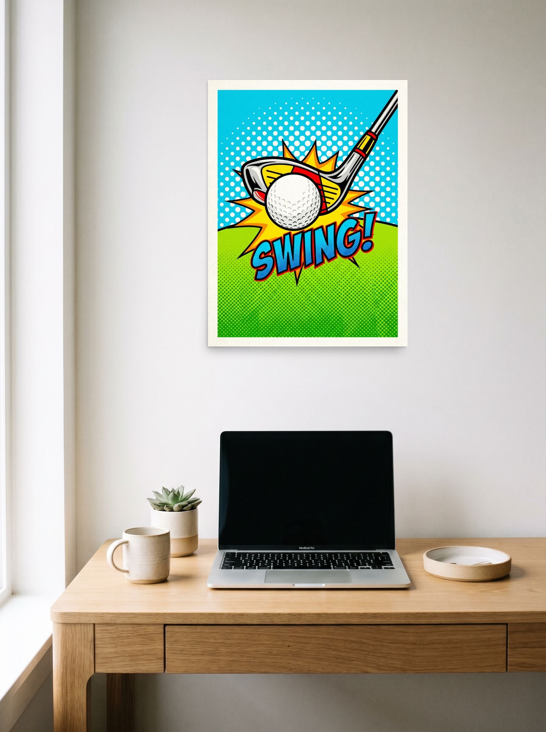A 18x24 inch unframed paper Pop art print of 'Comic Swing' hanging above a worktable with a laptop.