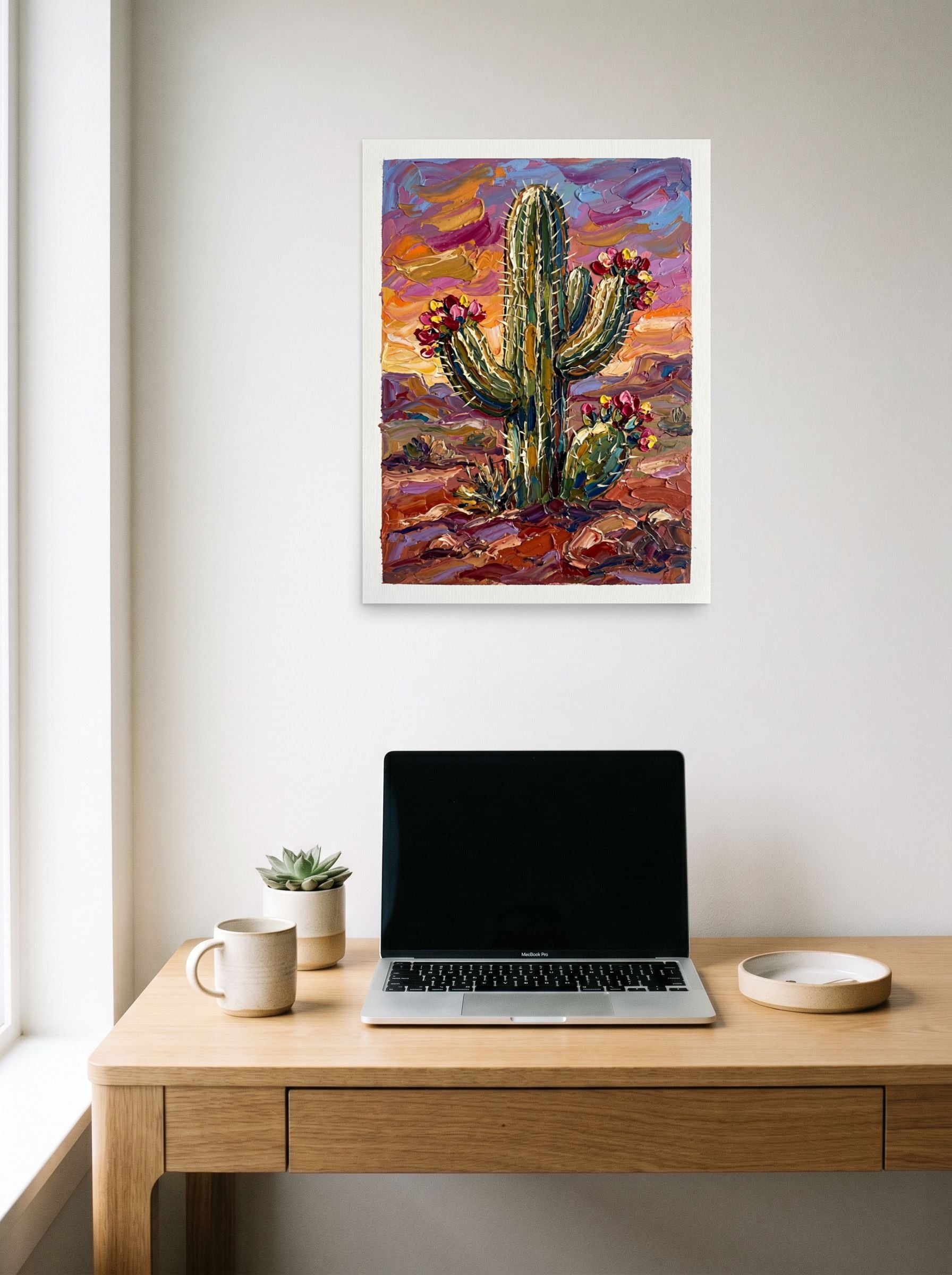 A 18x24 inch unframed paper Expressionism print of 'Desert Bloom' hanging above a worktable with a laptop.
