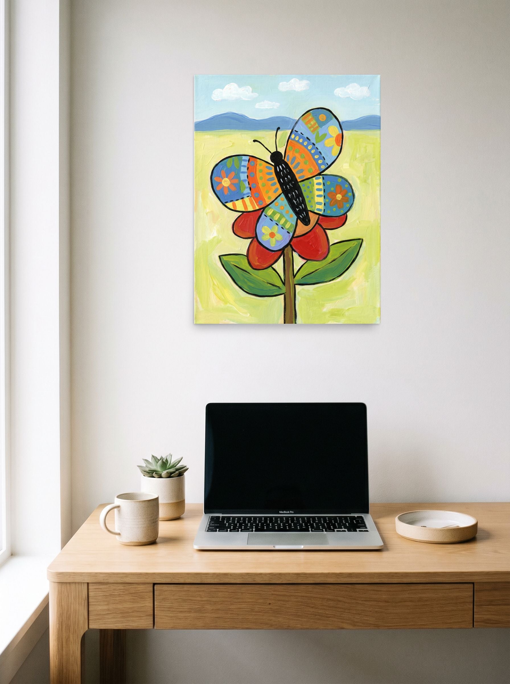 A 18x24 inch unframed paper Naive art print of 'The Patchwork Butterfly' hanging above a worktable with a laptop.