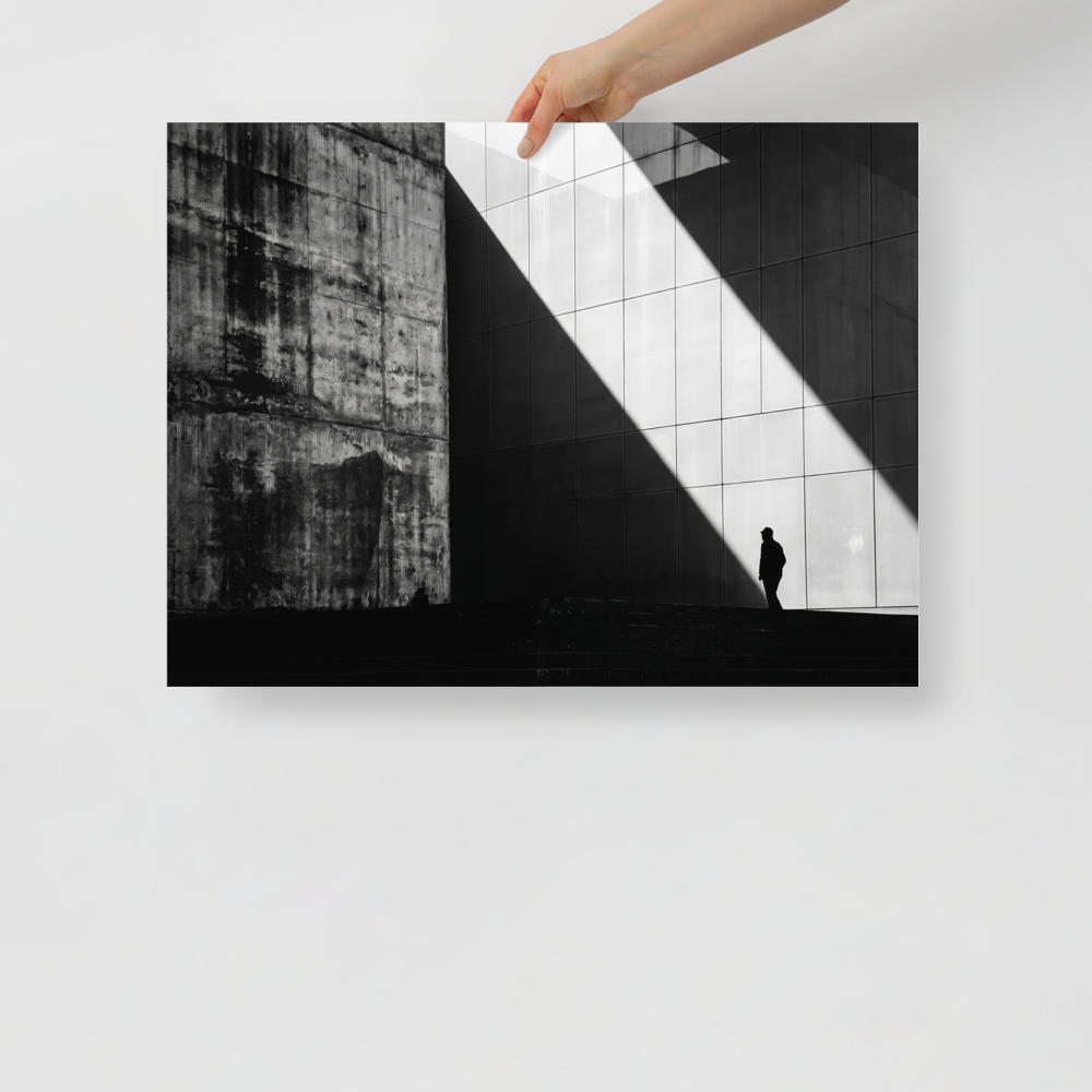 'Urban Geometry' — a Modernism print. A high-contrast black and white architectural photograph featuring a sharp diagonal shaft of light and a lone silhouette walking past textured concrete walls, perfect for an Office or Modern Living Room.