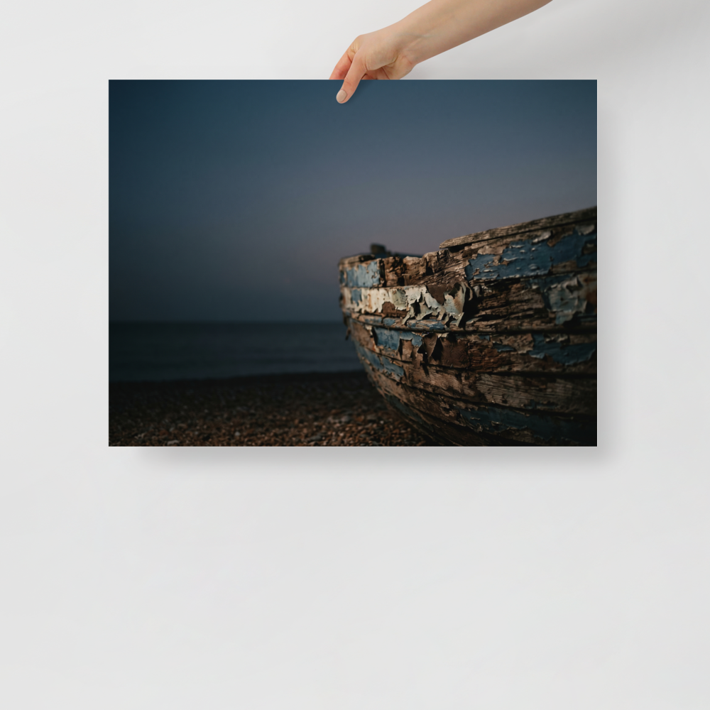 'The Faded Blue Boat' — a Art photography print. Close-up photograph of a weathered wooden boat with peeling blue paint resting on a rocky beach at dusk, suitable for a serene bathroom or living room.