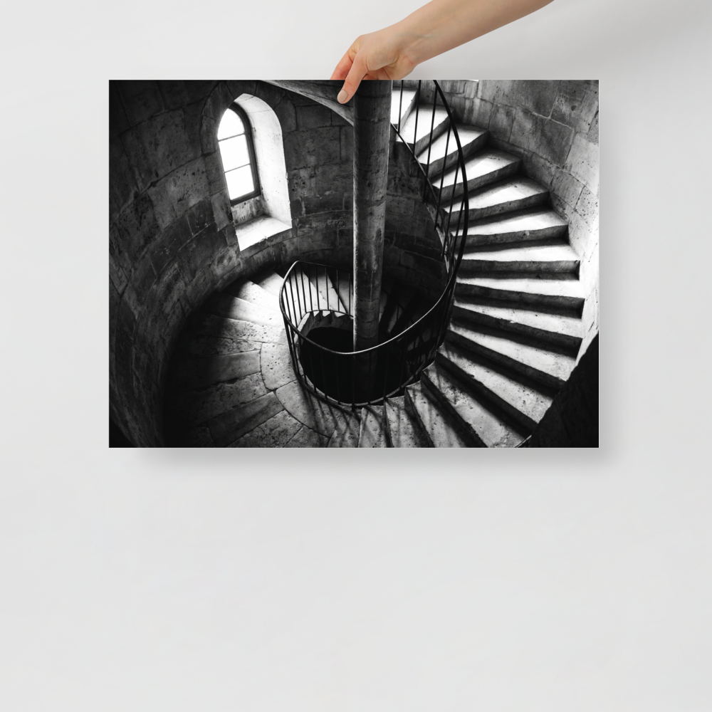 'Descent into Light' — a Art photography print. A high-contrast black and white photograph looking down a spiral stone staircase illuminated by a small window, adding depth and intrigue to an Office or Hallway.
