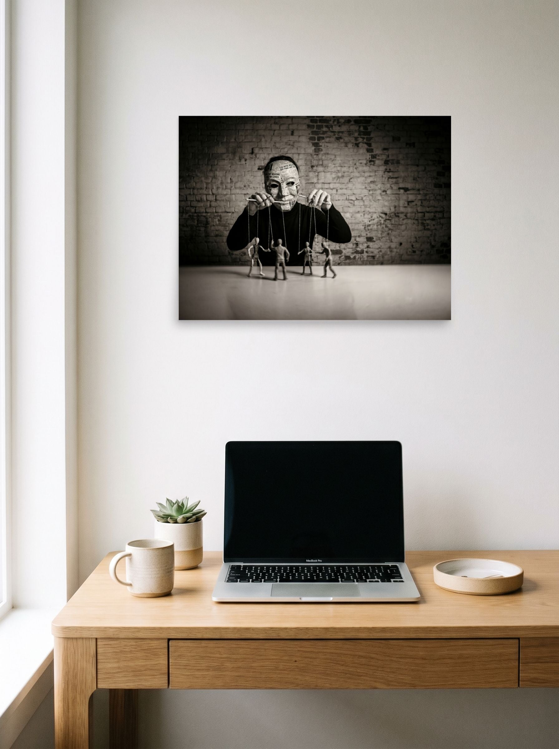 A 24x18 inch unframed paper Surrealism print of 'The Puppeteer of News' hanging above a worktable with a laptop.