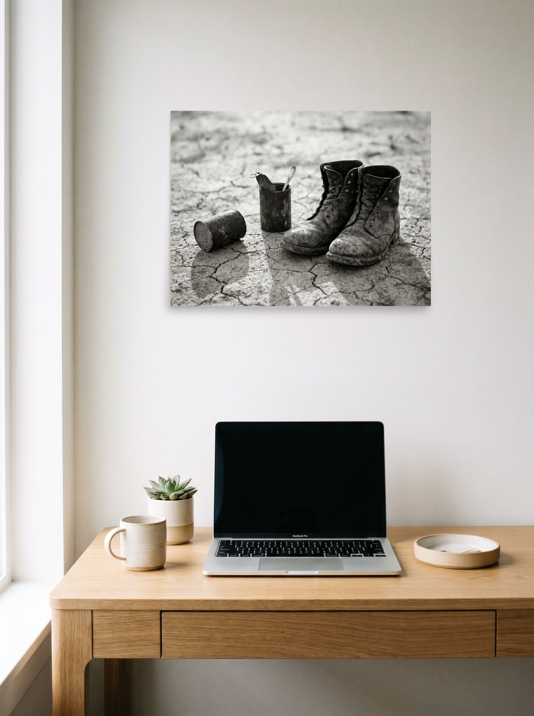 A 24x18 inch unframed paper American realism print of 'The Drifter's Boots' hanging above a worktable with a laptop.