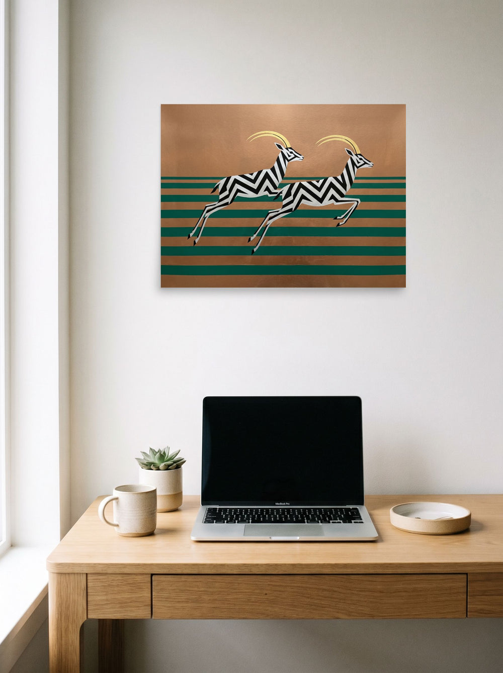 A 24x18 inch unframed paper Art Deco print of 'Golden Gazelles' hanging above a worktable with a laptop.