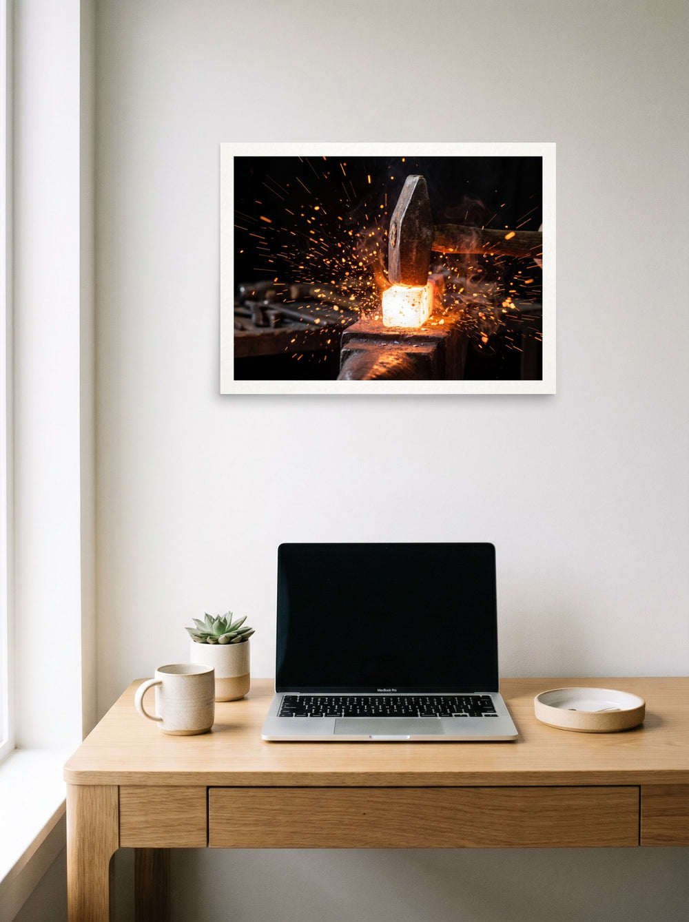 A 24x18 inch unframed paper Art photography print of 'Strike of the Anvil' hanging above a worktable with a laptop.