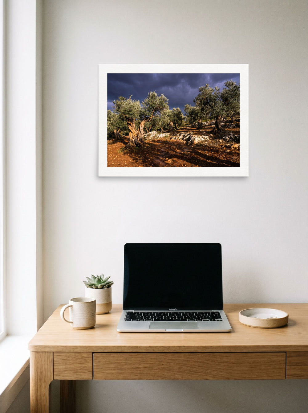 A 24x18 inch unframed paper Art photography print of 'Storm Over the Olive Grove' hanging above a worktable with a laptop.