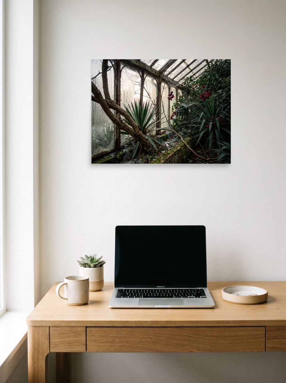 A 24x18 inch unframed paper Art photography print of 'The Overgrown Glasshouse' hanging above a worktable with a laptop.